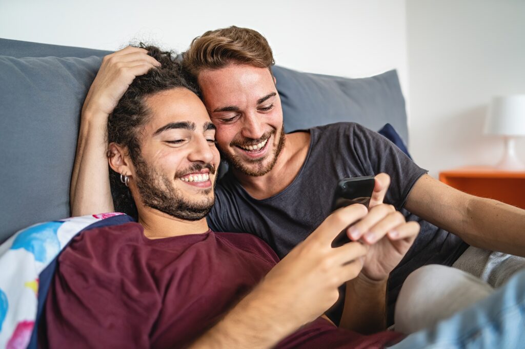 Affectionate gay couple watching content online with mobile cellphone in the bed