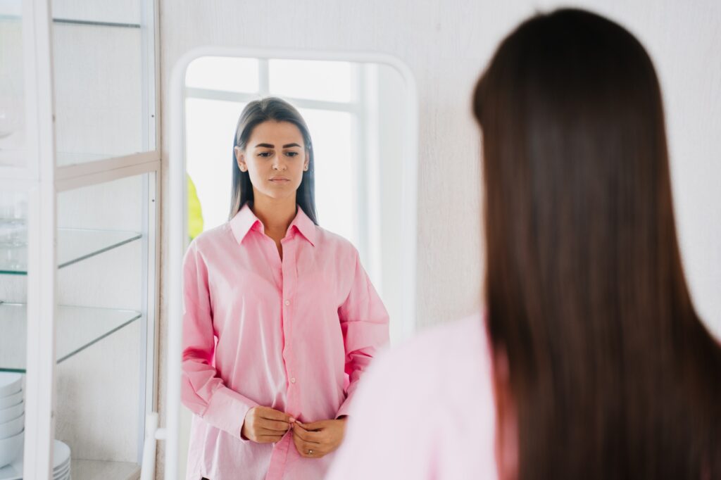 Sad brunette fit young woman in pink oversize shirt stands in front of mirror feels loneliness.